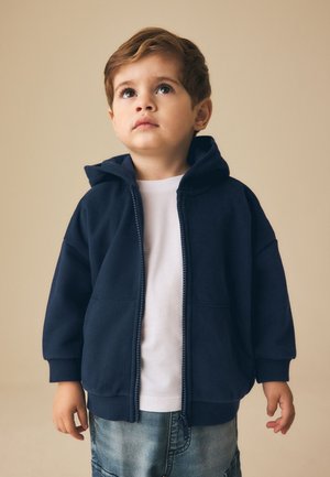 Junger Junge in navyblauem Hoodie über weißem Hemd und Jeans schaut vor beigem Hintergrund nach oben.