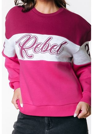 Kobieta nosząca bluzę typu color-block z ciemnoróżowymi, białymi i jasnoróżowymi paskami oraz napisem „Rebel” na piersi, napisanym kursywą.