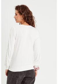 Top bianco a maniche lunghe con dettagli in pizzo sulle maniche, scollatura rotonda e bordo a coste. Modello che indossa jeans scuri, visto di spalle.