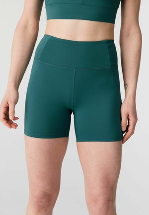 Femeie purtând pantaloni scurți atletici ajustați cu talie înaltă de culoare teal și un top scurt asortat, stând cu brațele relaxate pe lângă corp, pe un fundal gri.