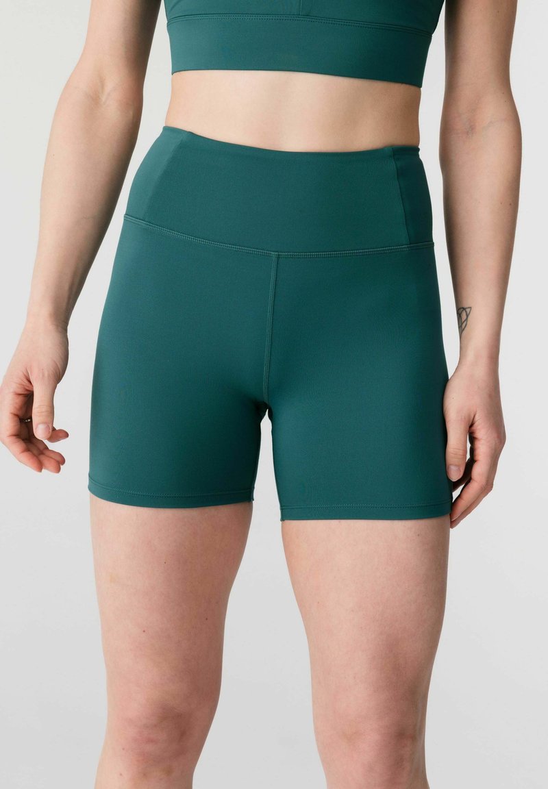 Naine, kes kannab rohelise tooni kõrge vöökohaga spordisorte ja sobivat lühikest toppi, seisab halli taustaga, käed lõdvestunult kõrval.