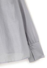 Blusa grigia chiaro con maniche lunghe, realizzata in un tessuto trasparente. Presenta polsini arrotondati con tre bottoni coperti in tessuto. Texture liscia.