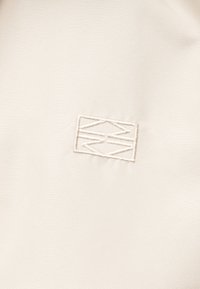 Logo géométrique brodé avec des lignes croisées à l'intérieur d'une bordure rectangulaire sur un fond de tissu beige lisse.