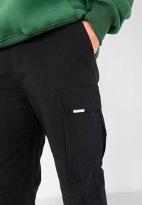 Zwarte cargobroek van duurzaam fabricage, met zijzakken en een klein merklabel aan de voorkant. Glad textuur, losse pasvorm.