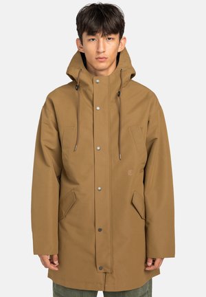 Junger Mann in einer braunen Kapuzenjacke mit Fronttaschen und Druckknöpfen, der vor einem einfachen hellen Hintergrund steht.