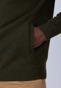 Pull en tricot vert foncé avec des poignets et un ourlet côtelés, doté d'une poche latérale. Le tissu semble doux avec une finition texturée.