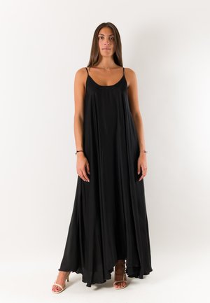 Donna in piedi che indossa un lungo vestito nero senza maniche con sottili spalline e sandali beige con tacco su sfondo bianco.