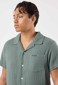 Camicia a maniche corte con bottoni, in tessuto verde smorzato e testurizzato, con una tasca sul petto e ricamo "SEASIDE" vicino alla tasca.