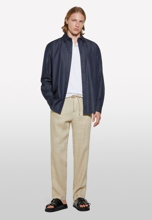 Chemise bleu marine à boutons sur un t-shirt blanc, associée à un pantalon en lin beige et des sandales noires. Comprend un col classique et une taille avec cordon de serrage.