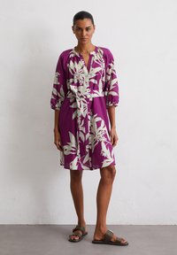 Paarse bloemenjurk met knoopsluiting, ceintuur in de taille en puffmouwen. De stof heeft een gladde textuur en heeft witte bladvormen.