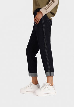 Jeans di denim scuro con cuciture chiare, orli risvoltati e una vestibilità rilassata, abbinati a sneaker bianche chunky con accenti grigi.