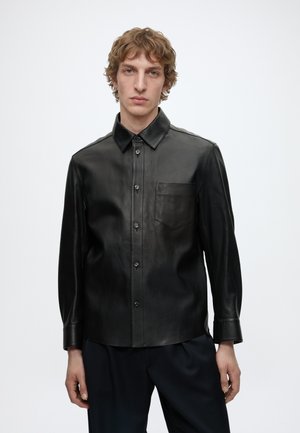 Jonge man met lichtbruin krullend haar, die een zwart leren overhemd met knopen en een donkere broek draagt, tegen een effen witte achtergrond.