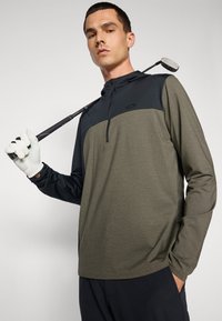 Langermet golfpullover i oliven grønn og marineblå, med glidelås foran og teksturert stoff, båret av en modell som holder en golfklubbe.