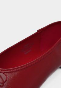 Κόκκινο δερμάτινο μπαλαρίνα με στρογγυλεμένη μύτη και μικρό φιόγκο, εμφανίζεται σε κοντινή γωνία σε λευκό φόντο.