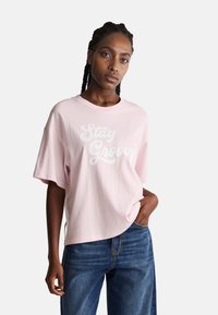 Růžové bavlněné tričko s nápisem "Stay Groovy" v bílé barvě, oversize střih, krátké rukávy, kulatý výstřih, kombinované s modrými denimovými džíny.