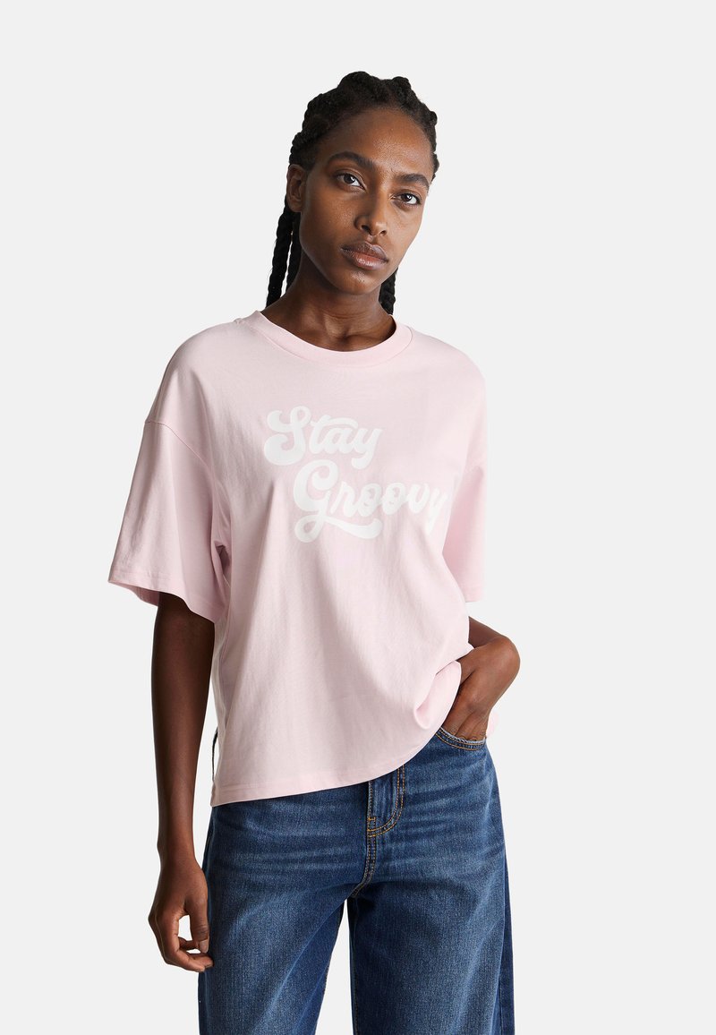 Růžové bavlněné tričko s nápisem "Stay Groovy" v bílé barvě, oversize střih, krátké rukávy, kulatý výstřih, kombinované s modrými denimovými džíny.