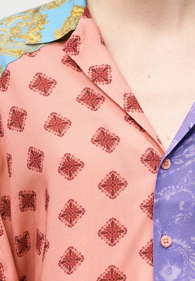 Πουκάμισο με κοντό μανίκι που διαθέτει ροζ σώμα με σκούρα κόκκινα λουλουδένια σχέδια, ανοιχτό μπλε γιακά με χρυσές λεπτομέρειες και πορφυρό μπροστινό μέρος με κουμπιά.