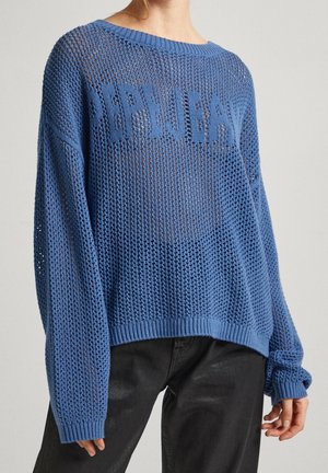 Femme portant un pull bleu ample en maille ouverte avec un col rond et un pantalon noir, debout devant un fond clair uni.
