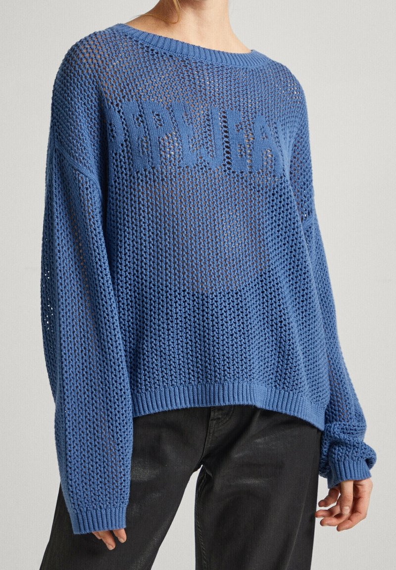 Femme portant un pull bleu ample en maille ouverte avec un col rond et un pantalon noir, debout devant un fond clair uni.
