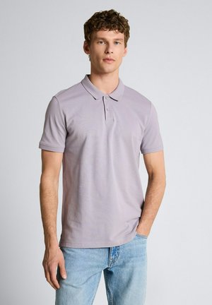 Jonge man met krullend haar, gekleed in een lichtpaars poloshirt en lichtblauwe spijkerbroek, staand tegen een effen grijze achtergrond.