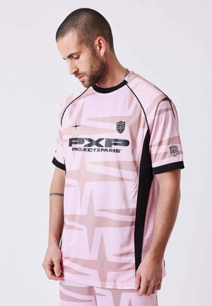 Uomo con capelli corti e barba che indossa una maglietta sportiva PXPD ProjectxParis rosa con motivi astratti, guarda verso il basso con le mani ai lati.