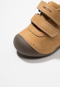 Scarpa scamosciata beige con puntale in gomma marrone, dotata di due cinturini in Velcro e cuciture bianche a contrasto per un tocco di stile.