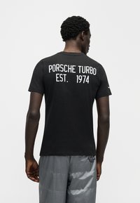 Μαύρο κοντομάνικο μπλουζάκι με λευκό κείμενο "PORSCHE TURBO EST. 1974" στην πλάτη. Ομαλή ύφανση, κλασικός σχεδιασμός με στρογγυλή λαιμόκοψη.