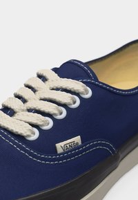 Κοντινή λήψη ενός μπλε αθλητικού παπουτσιού Vans με λευκά κορδόνια, λευκές οπές, εμφανείς ραφές και μπεζ εσωτερική επένδυση.