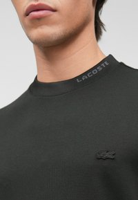 Μαύρη μπλούζα με ριχτό λαιμόκοψη. Διαθέτει ορατό λογότυπο Lacoste σε συμβατό χρώμα στον γιακά και ένα μικρό κεντημένο λογότυπο στο στήθος.