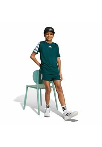 Jongen in teal sportoutfit en zwarte pet leunt tegen een turquoise stoel, draagt witte sneakers en gestreepte sokken op een witte achtergrond.