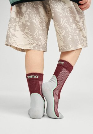 Child wearing beige patterned shorts and maroon-gray socks standing on tiptoes on a light background.