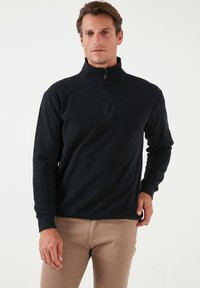 Pullover in pile nero con colletto alto e chiusura a zip metà. Presenta maniche lunghe e una texture liscia e morbida. Il design minimalista è funzionale.