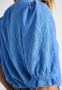 Blå texturerad tygblus med puffiga korta ärmar och intrikata hålmönster i diamantform, bärs av en person.