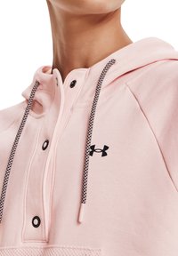 Sudadera con capucha rosa y cuello abotonado, con detalles en negro y un logo. Presenta tela texturizada y cordones de contraste.