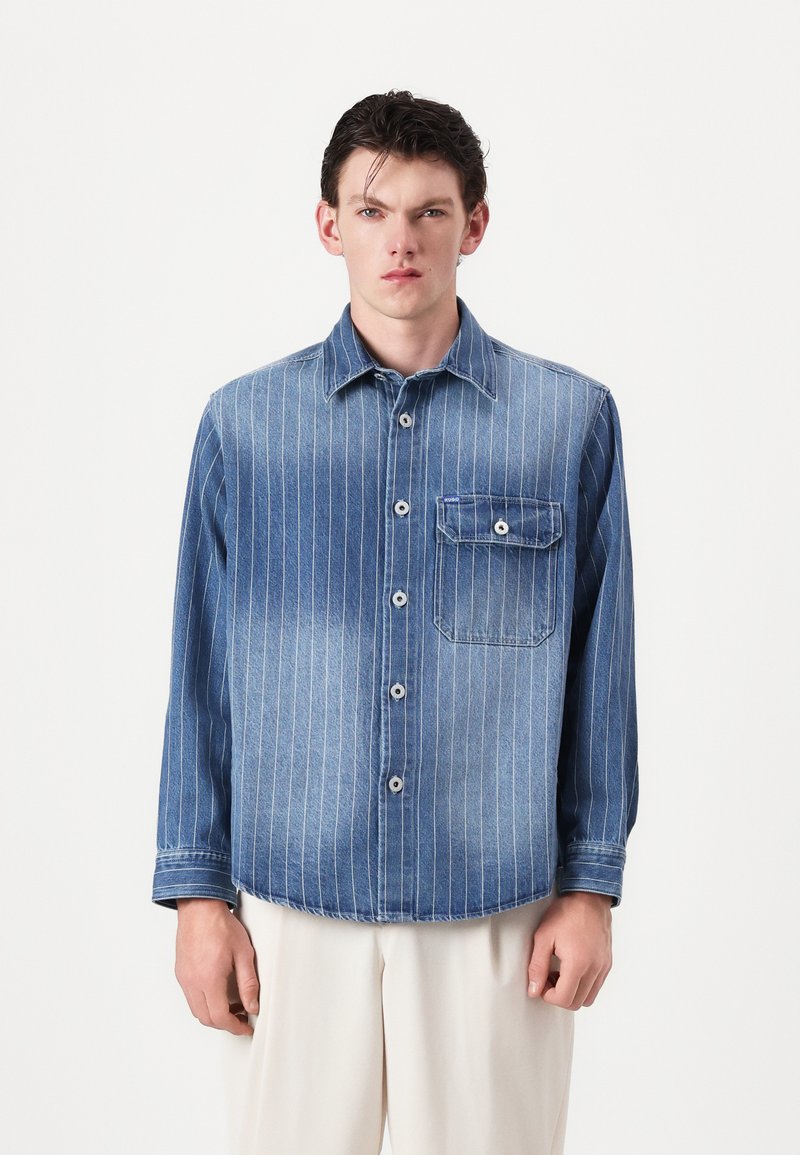 Denim shirt featuring vertical white stripes, a front pocket, and button closure. Light blue color with a smooth texture and relaxed fit.