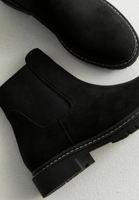 Black suede ankle boots with elastic side panels, round toe, and contrasting white stitching on a thick sole. Smooth texture and sturdy design.