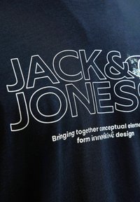 Tamsiai mėlyna marškinė su baltu spausdintu tekstu. Yra prekės ženklas "JACK & JONES" ir šūkio detalės, pateiktos skirtingais šrifto dydžiais. Pagaminta iš medvilnės.