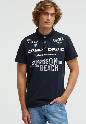 Junger Mann mit lockigem Haar, der ein marineblaues Polohemd mit weißem, strandbezogenem Schriftzug und ausgewaschene blaue Jeans trägt, eine Hand in der Tasche.