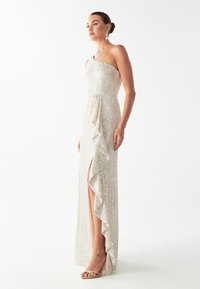 Vestido de noche de un solo hombro, con lentejuelas en color beige claro, con una abertura alta en el muslo y detalle de volantes. Combinado con sandalias de tiras nude y pendientes.