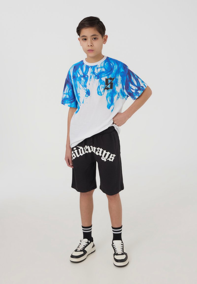 Ragazzo in piedi con una mano sui fianchi, indossa una camicia bianca con motivo a fiamme blu, pantaloni neri con testo "sideways", calze a righe e scarpe bianche e nere.