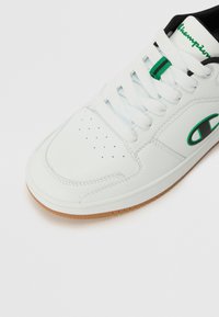 Sneaker din piele albă cu perforații pe vârf, accente verzi și negre, și o talpă din cauciuc bej. Sireturile sunt albe, având detalii mici verzi.