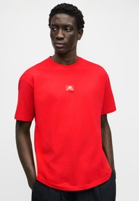 Άνδρας με κοντά σγουρά μαλλιά, φορώντας ένα φωτεινό κόκκινο μπλουζάκι με ένα μικρό λευκό γραφικό στο κέντρο του στήθους, στέκεται μπροστά σε ένα απλό φόντο.