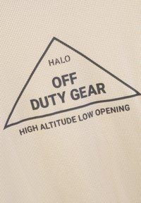 Ecru materiał o fakturze, z czarnym trójkątnym nadrukiem i napisem "HALO OFF DUTY GEAR WYSOKA WYSOKOŚĆ NISKIE OTWARCIE".