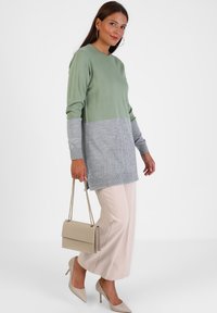 Hellgrüner und grauer Pullover mit langen Ärmeln, der einen strukturierten grauen unteren Abschnitt und gerippte Bündchen aufweist. Kombiniert mit beigen, weiten Hosen.