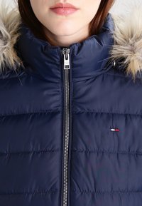 Navy blue puffer jacket with a zip closure, featuring a fur-lined hood and horizontal quilted pattern. Small logo accent near the hem.