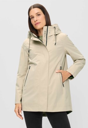 Frau trägt einen beigen Regenmantel mit Kapuze und Reißverschlusstaschen, eine Hand in der Tasche, posiert vor einem hellgrauen Hintergrund.