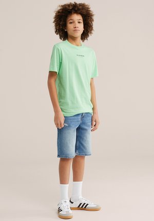Niño con cabello rizado que lleva una camiseta verde claro con el texto "Team Blue", pantalones cortos de mezclilla, calcetines blancos y zapatillas Adidas blancas con rayas negras.