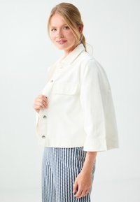 Jeune femme aux cheveux blonds en queue de cheval, portant une veste blanche boutonnée et un pantalon à motifs bleu et blanc sur un fond uni.