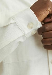 Camisa de botones de color beige claro con una tela texturizada. El puño presenta dos botones y una costura recta y ordenada. Patrón sutil visible.