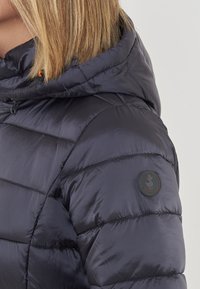 Donna che indossa una giacca imbottita di un blu navy scuro con cappuccio e una toppa rotonda con il logo "Save the Duck" sul braccio superiore.